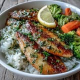 Golden herb-baked tilapia fillets resting on fluffy white rice with steamed broccoli and carrots.