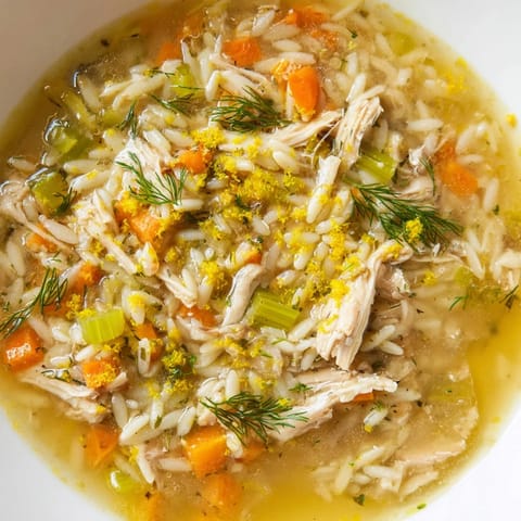 Tender chicken and orzo pasta swim in a golden, lemon-infused broth with diced carrots and celery in a rustic bowl.